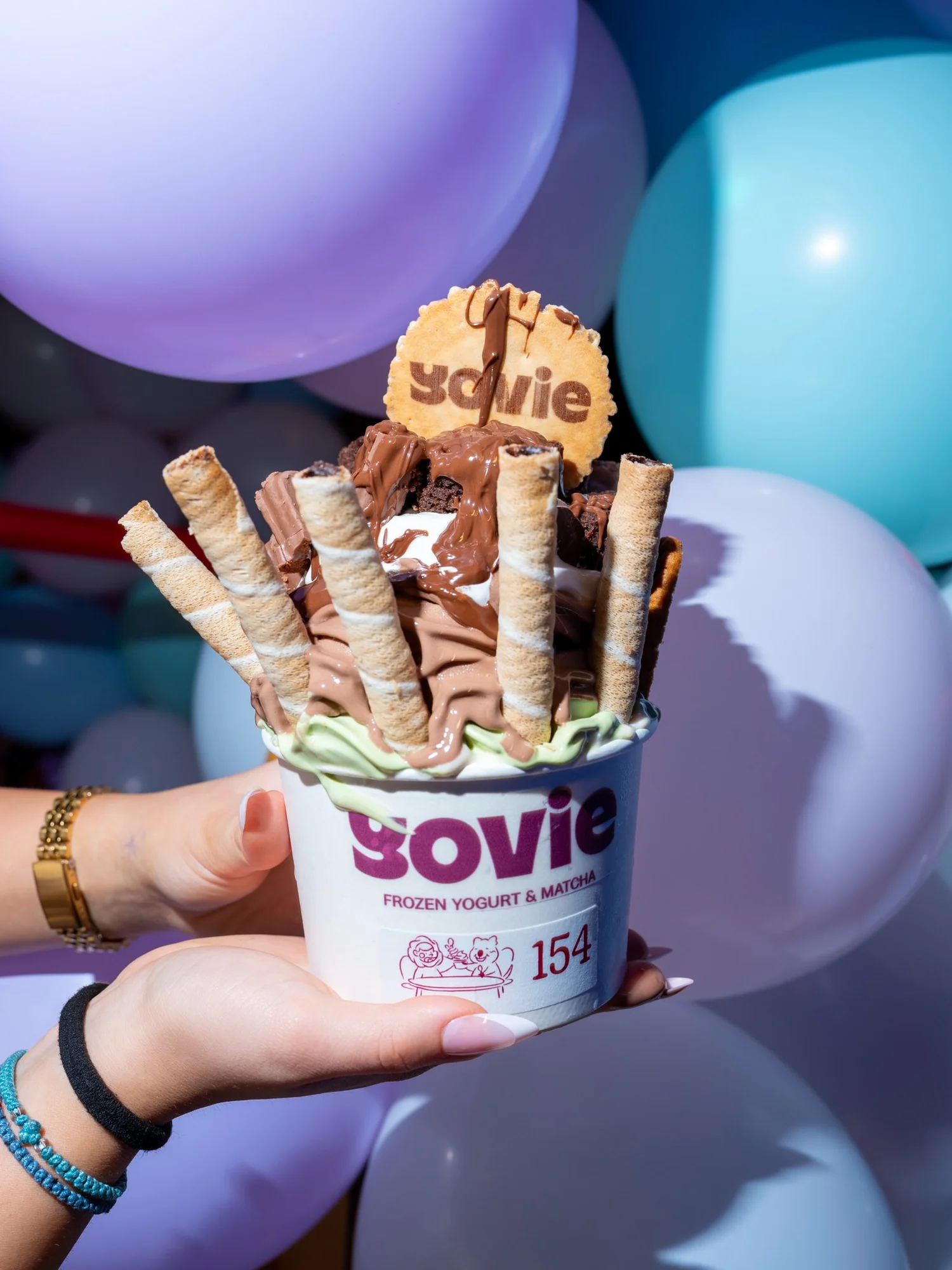 Yovie frozen yoghurt with chocolate, wafers and a branded biscuit against pastel balloons