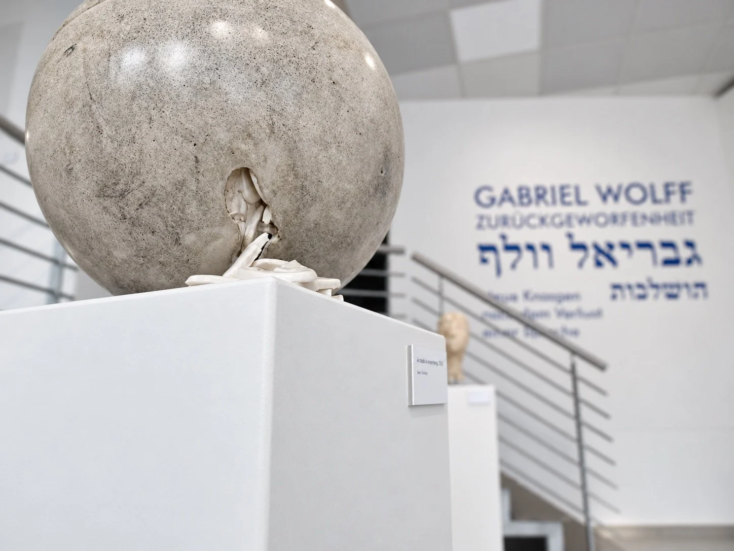 A concrete sphere with a crack, on a white plinth in the foreground; the blue title wall and the stairway behind.