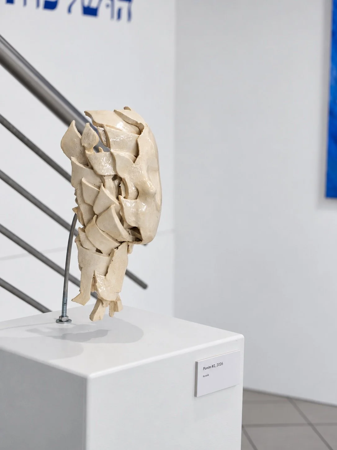 A ceramic head made of fragmented pieces, mounted on a thin metal stem on a white plinth in front of the title wall, with a stair railing at left.