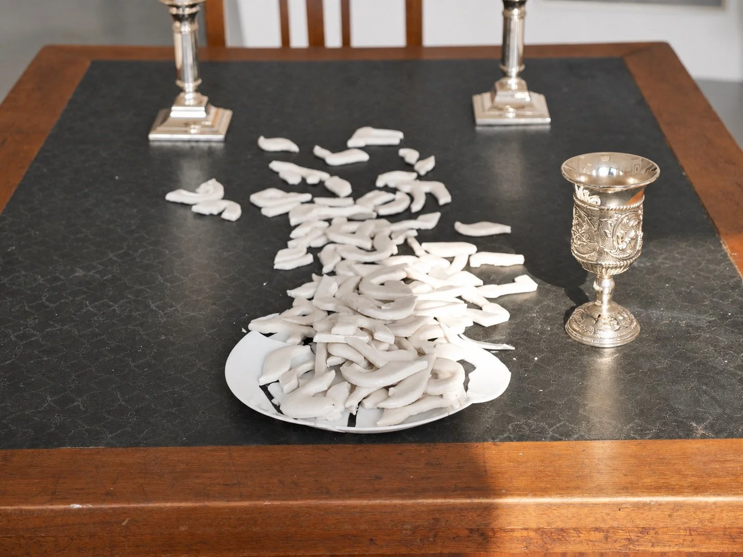 A dark-topped wooden table with a plate of broken white porcelain letters, a silver Kiddush cup and two candlesticks.