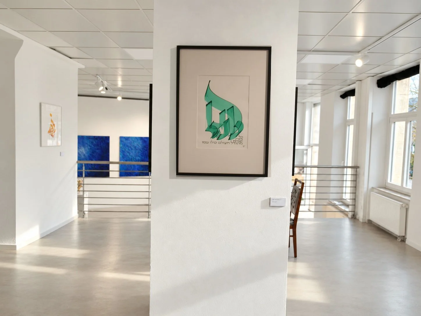 A framed green calligraphic work on a white column; blue paintings, a window and a radiator in the background.