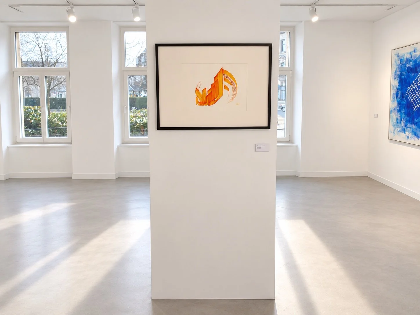 A framed orange calligraphic work on a freestanding wall between tall windows, with light falling across the floor.