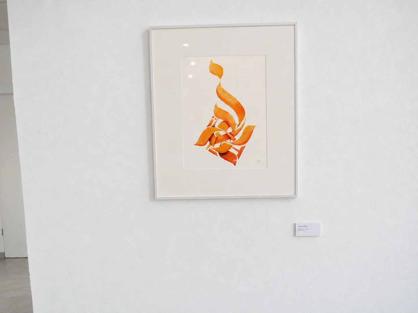 A framed orange calligraphic work with a white frame on a white wall, with a small label beside it.