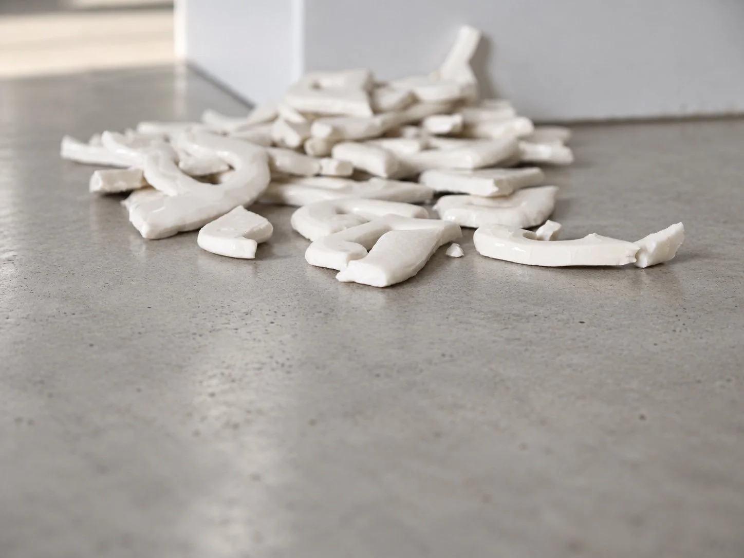 Pieces of broken white porcelain letters scattered on a concrete floor, with a white wall behind.
