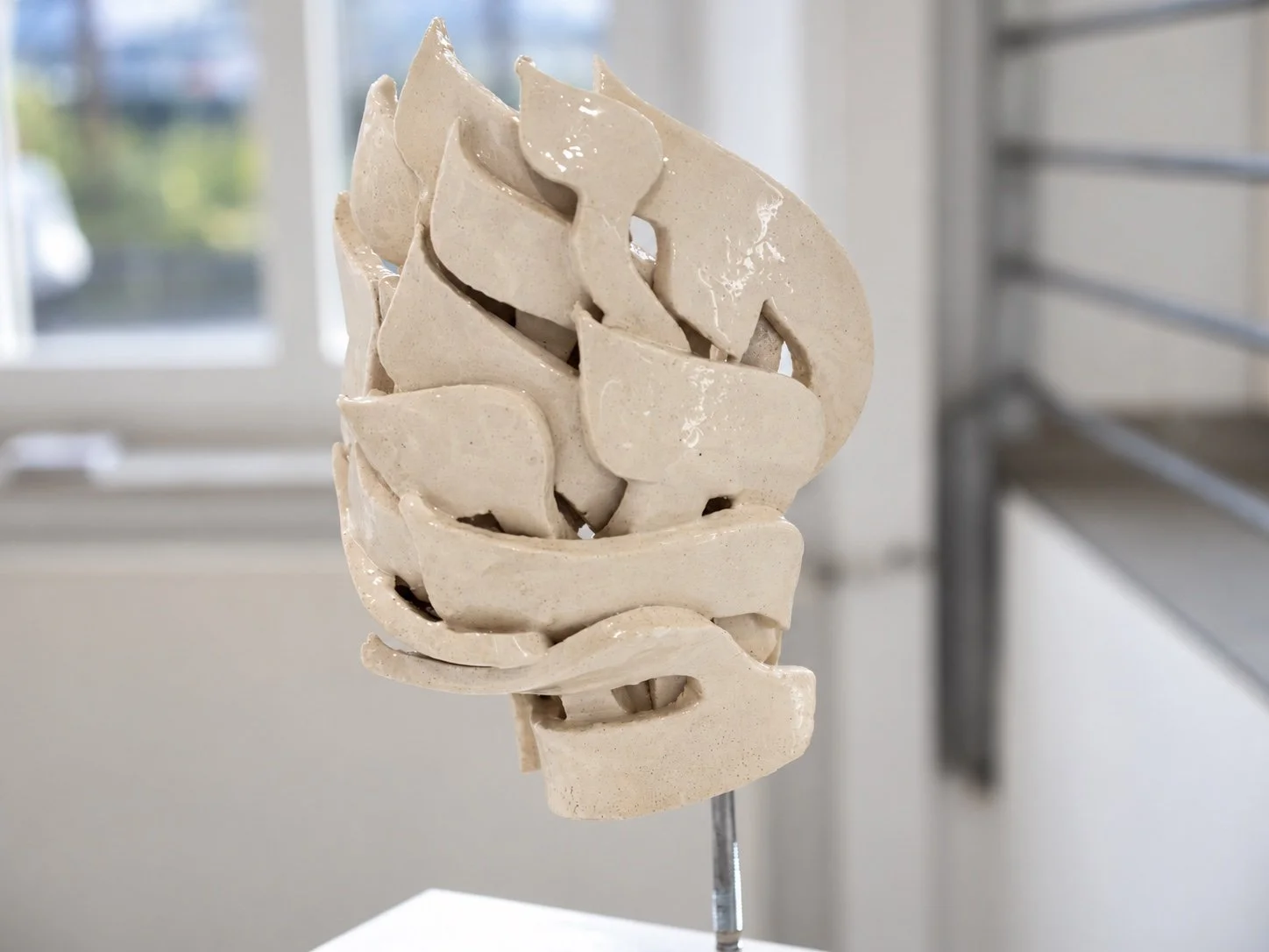 Close view of a cream-coloured ceramic sculpture built from layered leaf-like Hebrew letter forms, with a window behind.