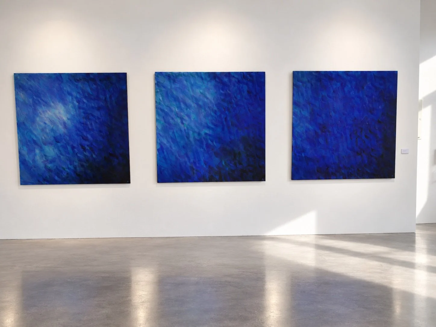Three square blue paintings in a row on a white wall above a concrete floor.