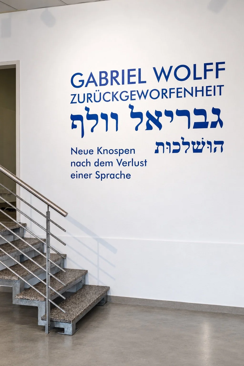 White entrance wall with the blue exhibition title in German and Hebrew beside a stairway with a steel railing.