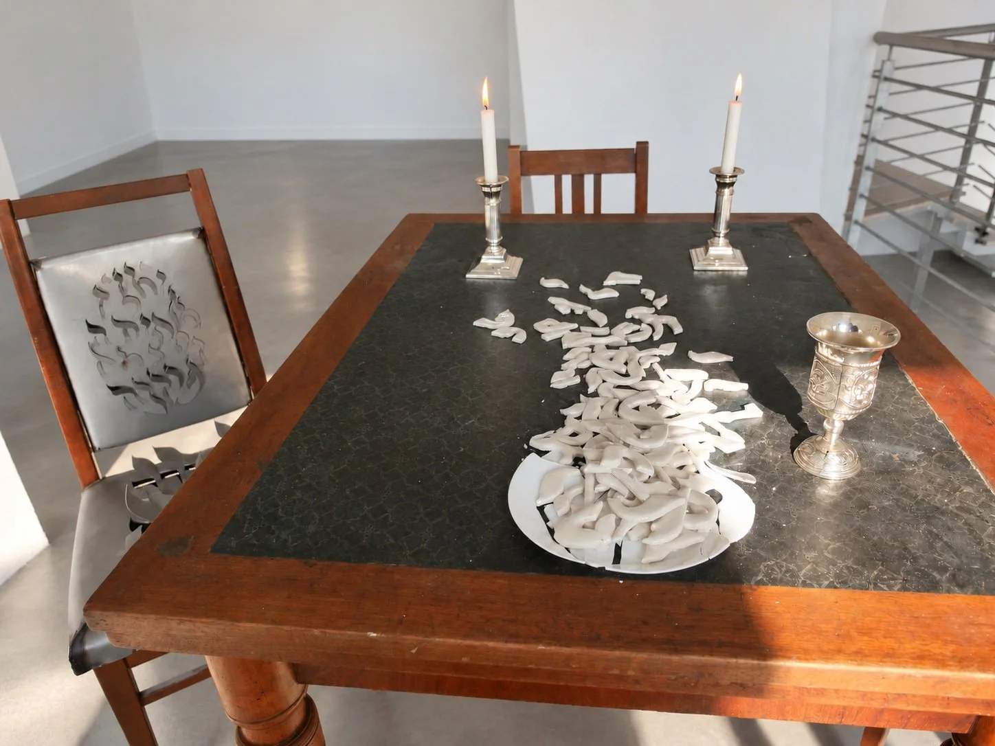 A wooden table with two lit candles in silver candlesticks, a plate of porcelain fragments and a chair with a steel seat at left.
