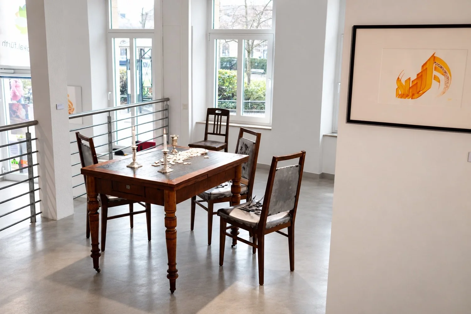 A wooden table with chairs near tall windows and a stair railing; a framed orange work hangs at right. Candles and objects stand on the table.