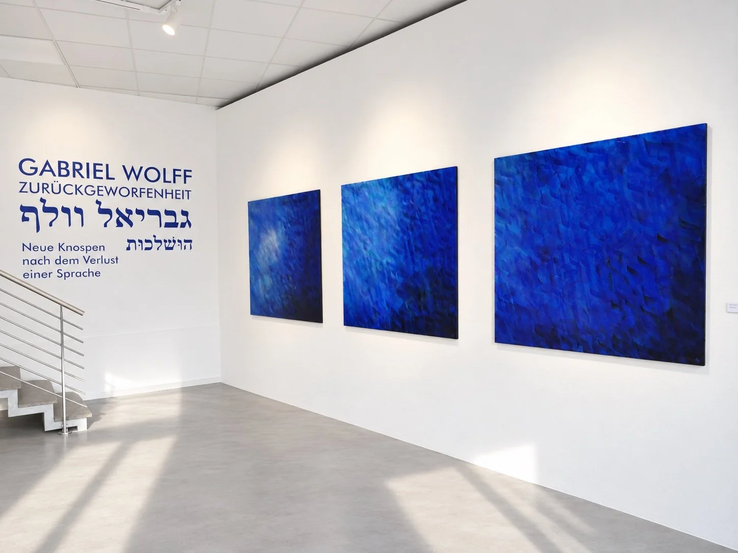 Three large blue paintings hang on a white wall to the right of the title wall; sunlight crosses the concrete floor.