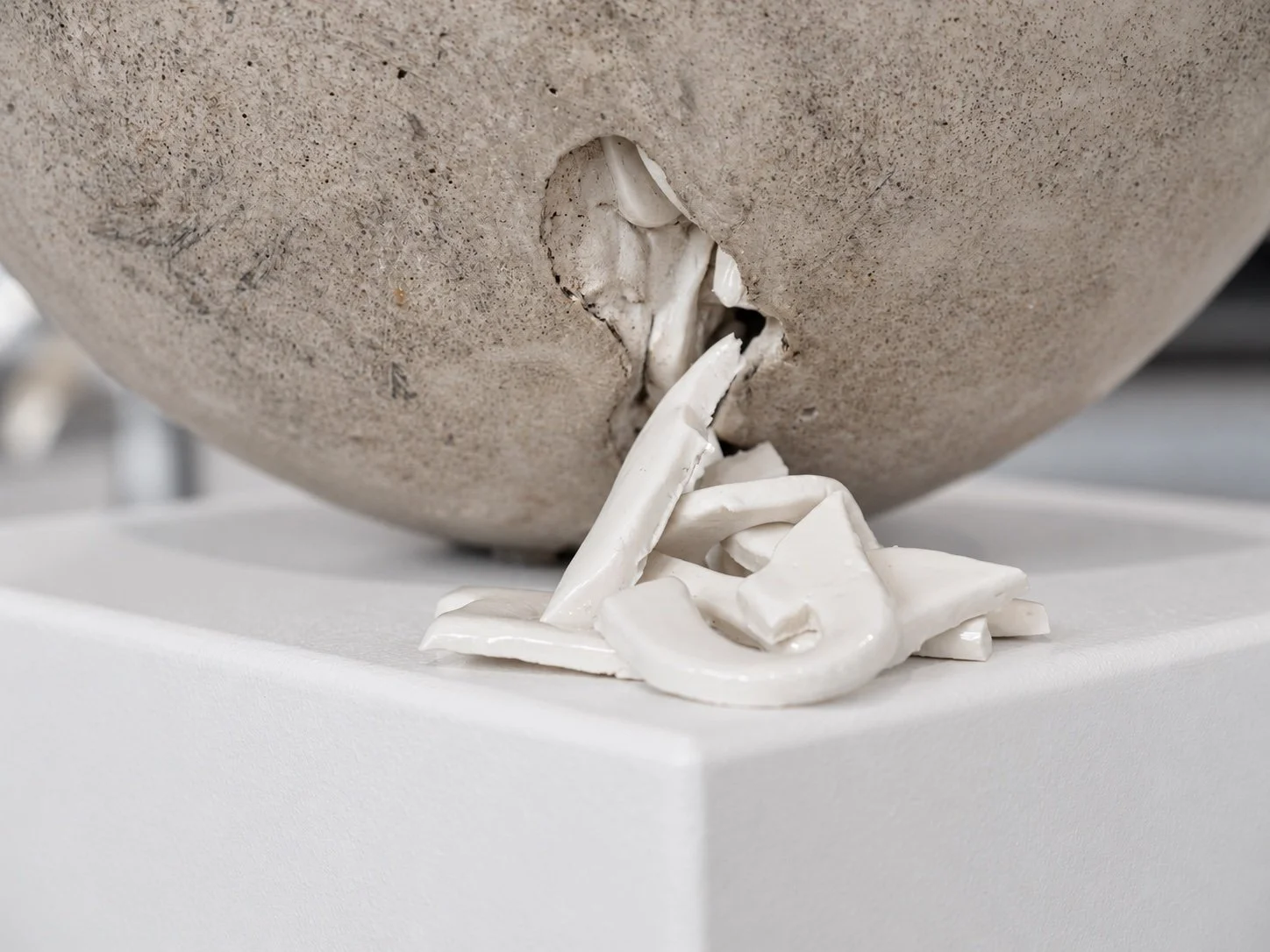 Close view of a crack in the concrete sphere, with white porcelain letter fragments spilling out onto the plinth.