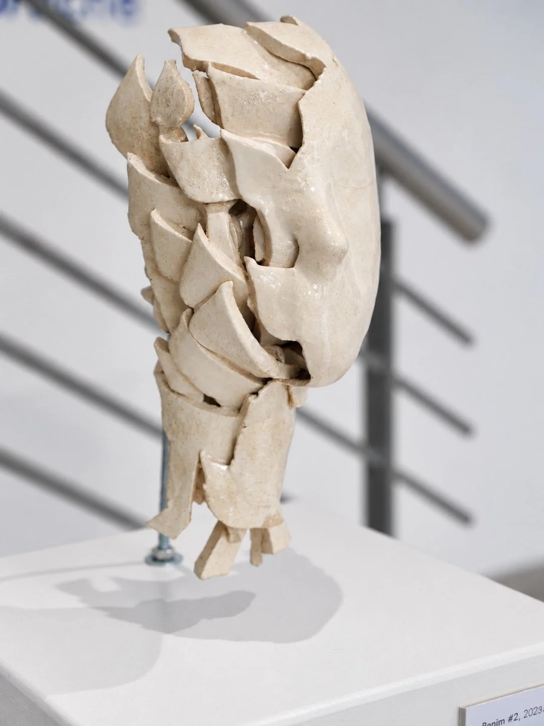 Side view of a ceramic head built from layered, broken fragments, on a white plinth with a stair railing behind.