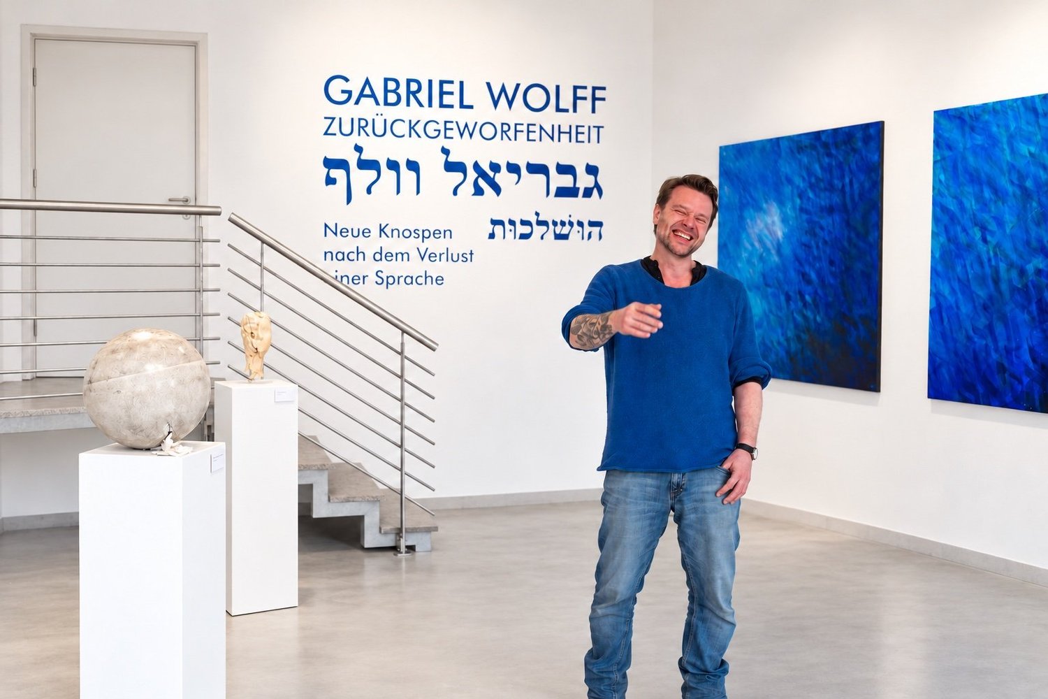 Gabriel Wolff, laughing, stands in the exhibition space in front of the title wall, with blue paintings at right and plinths at left.