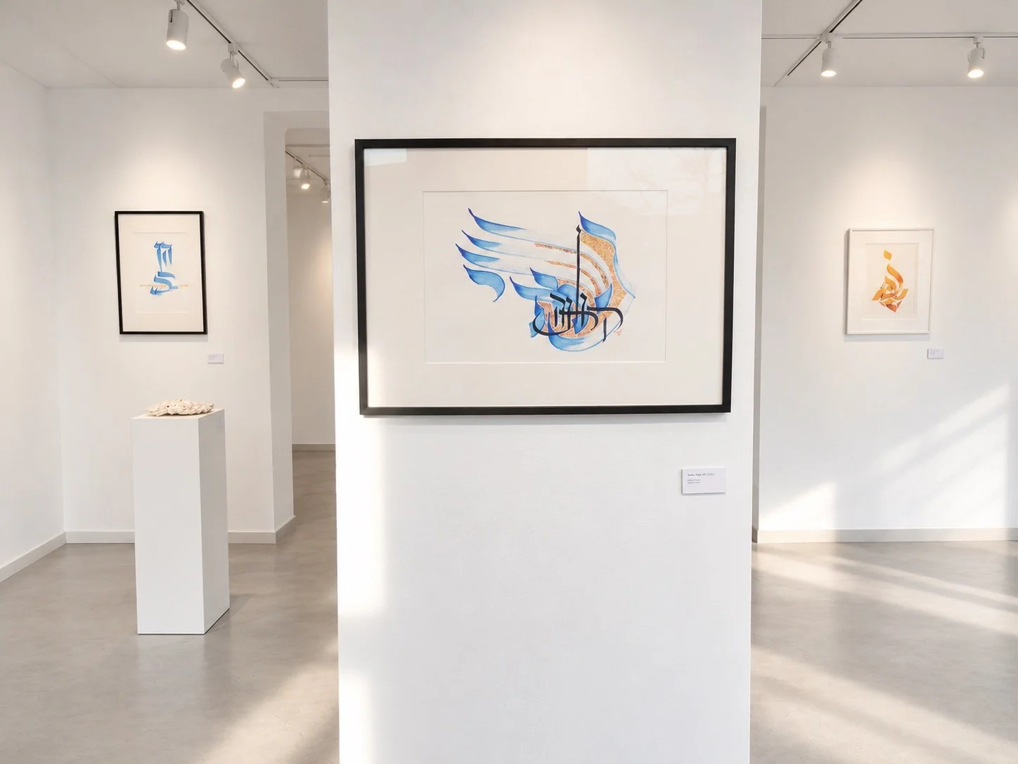 Gallery view with two framed calligraphic works on a white column, a wooden table with chairs and blue paintings in the distance.