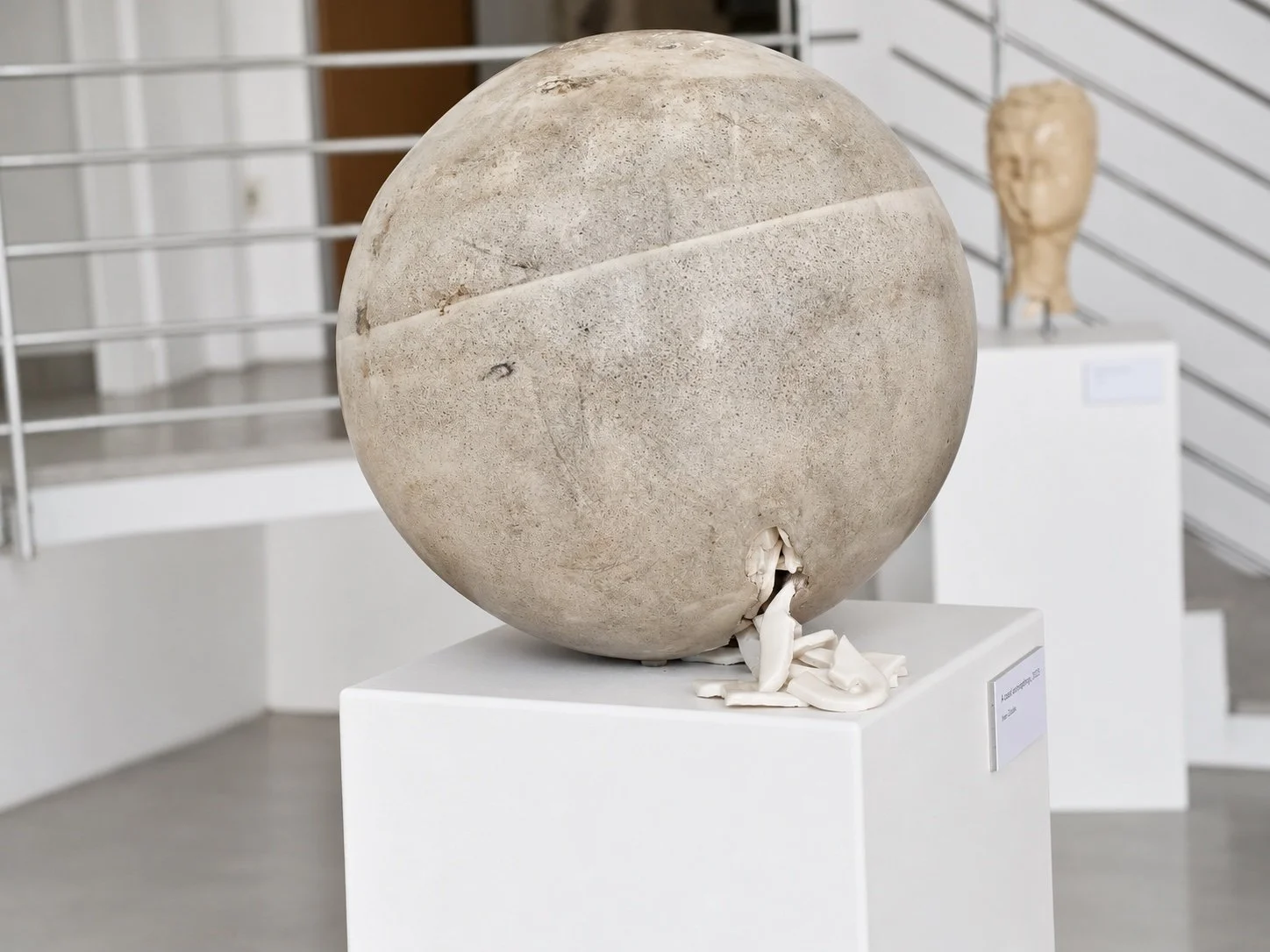 A concrete sphere with a horizontal seam across its surface and a crack at the lower edge, on a white plinth; a ceramic head on a plinth behind.
