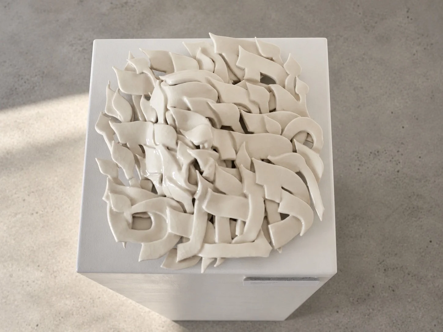 The porcelain sculpture seen from above, a dense relief of overlapping white Hebrew letter forms on a white plinth.