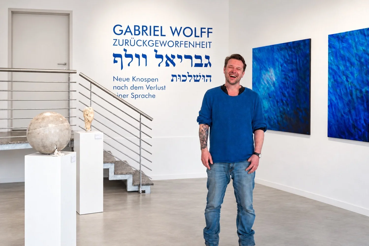 Gabriel Wolff, smiling, stands in the exhibition space in front of the blue title wall. Two blue paintings hang at right and sculptures on white plinths stand at left.