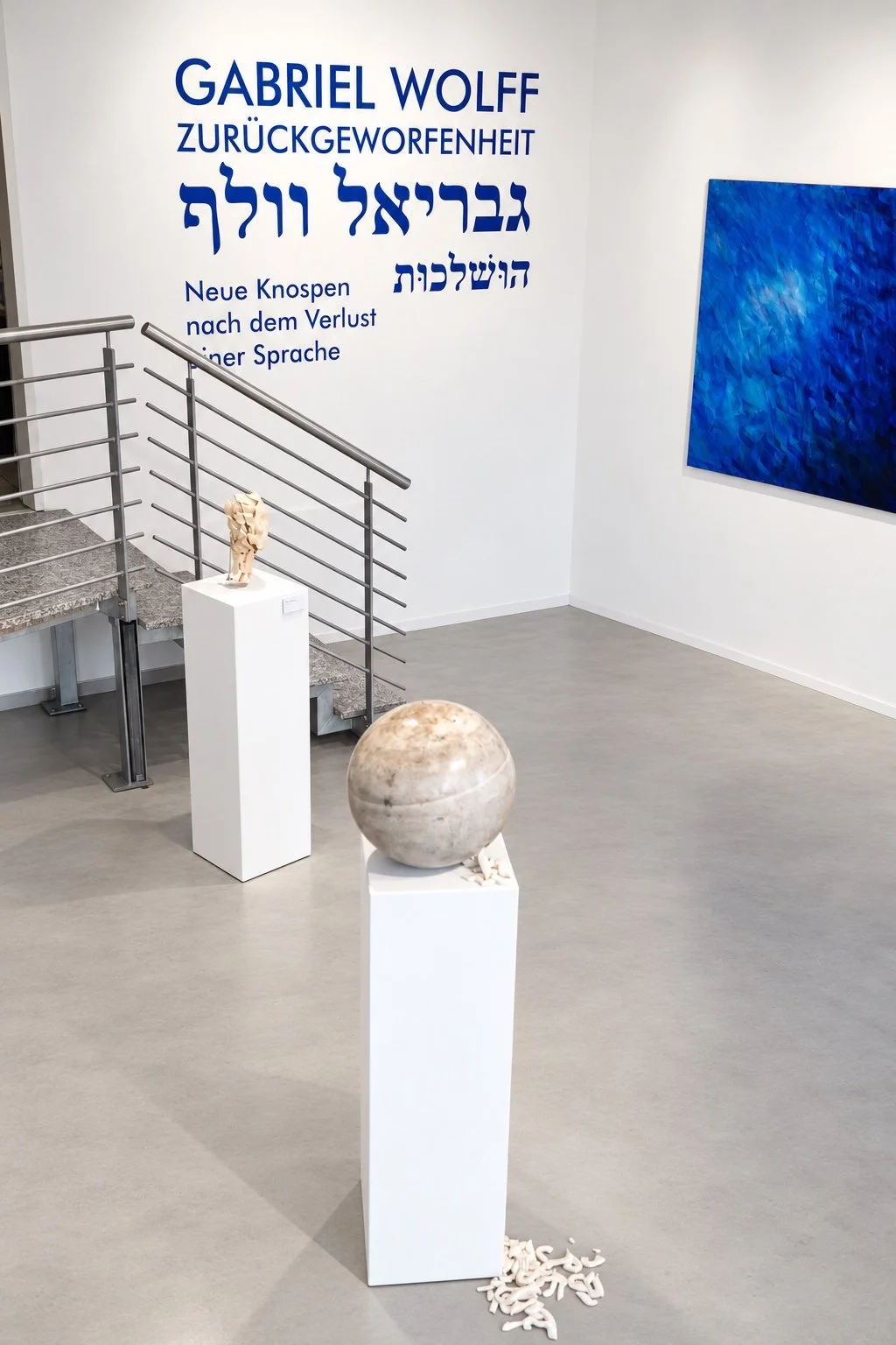 View towards the title wall: a concrete sphere and a ceramic head on white plinths in front of the stairway, with a blue painting at right.