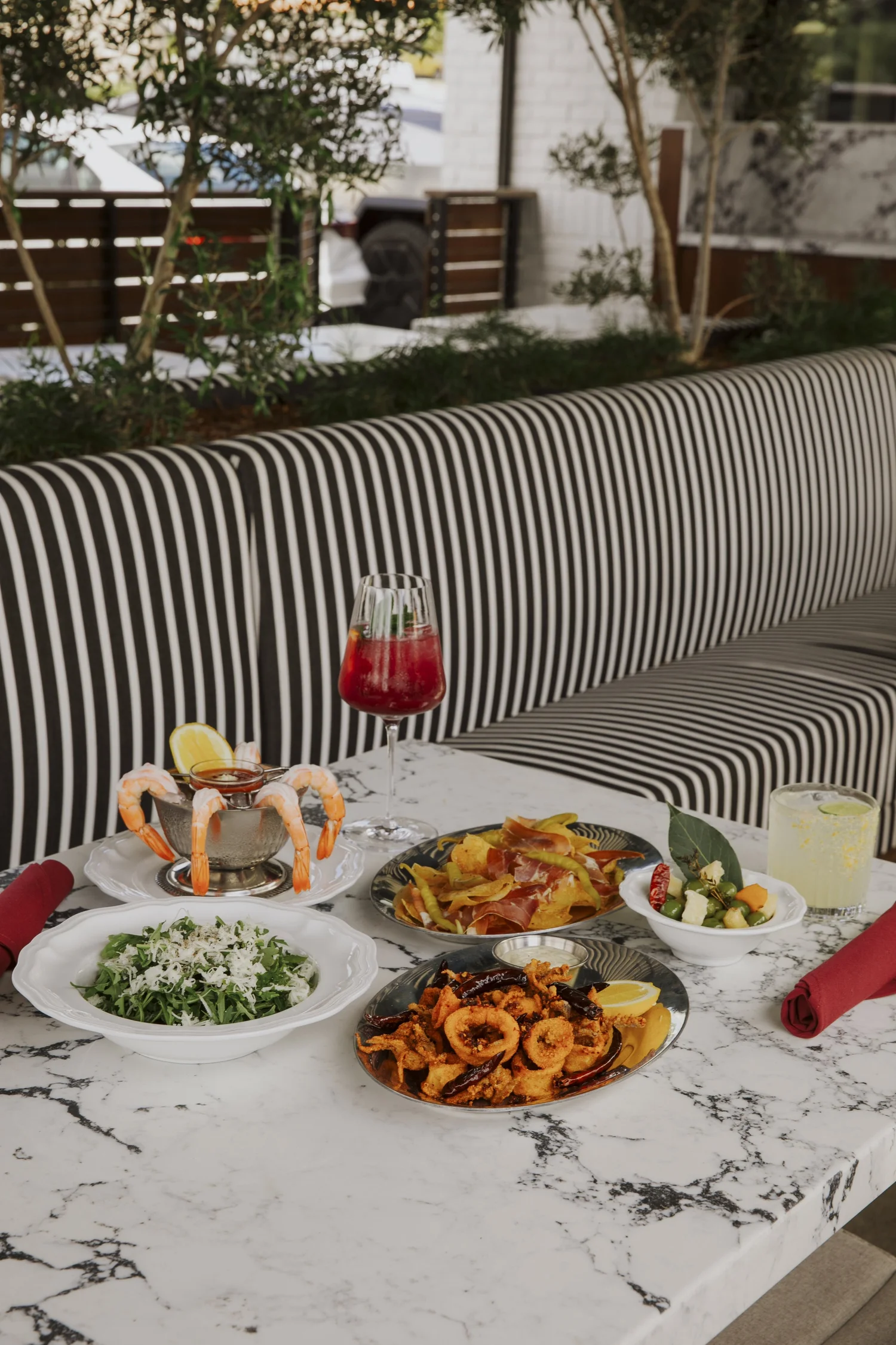 Salina's patio with a striped banquette under olive trees, set with shrimp, calamari and cocktails