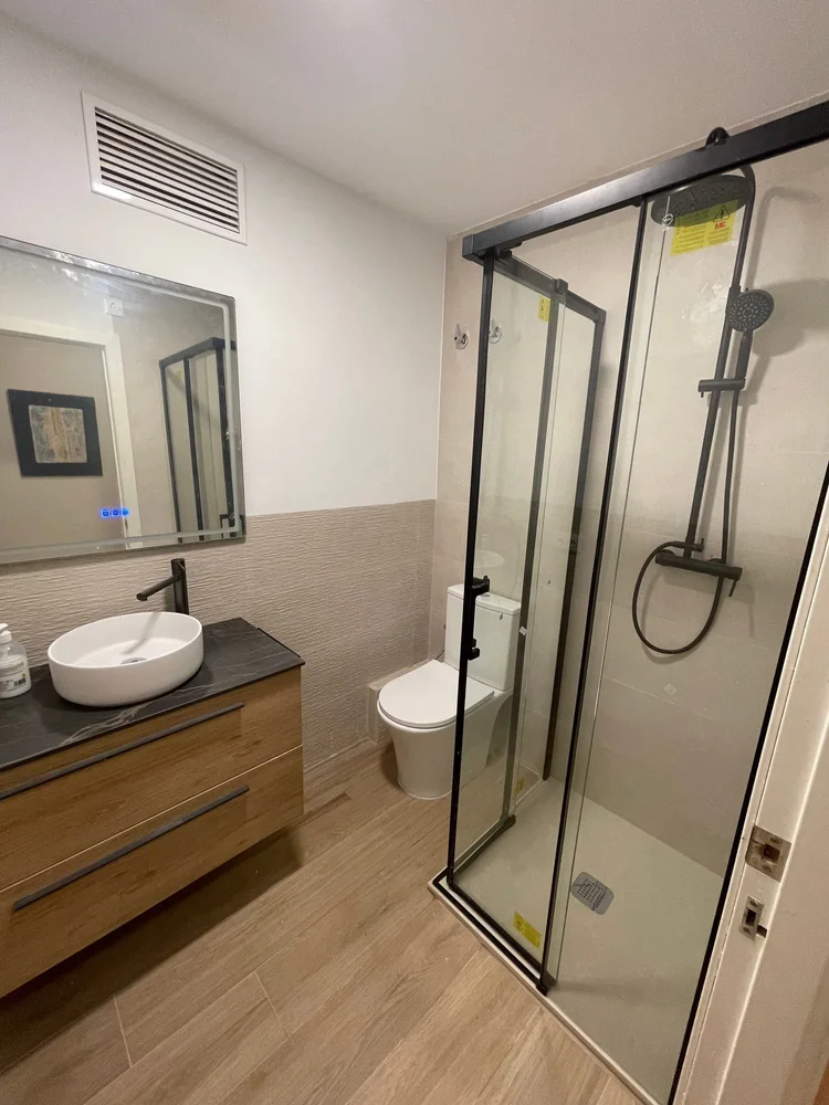 Baño con mueble de madera, lavabo sobre encimera y mampara de perfil negro