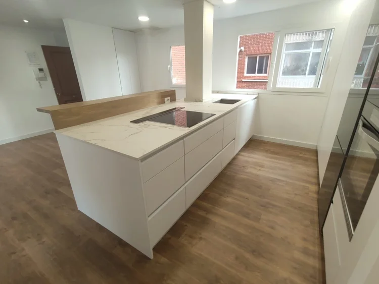 Cocina blanca reformada con península de encimera efecto mármol y barra de madera