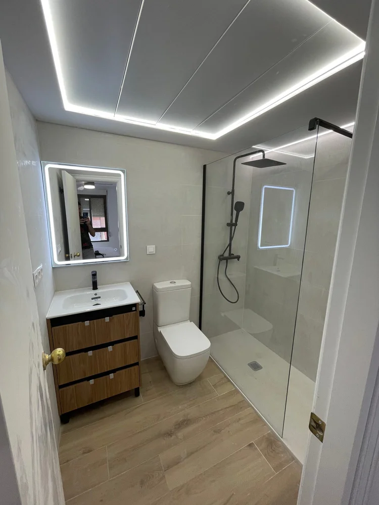 Baño reformado con techo de LED perimetral, mueble de madera y mampara de cristal