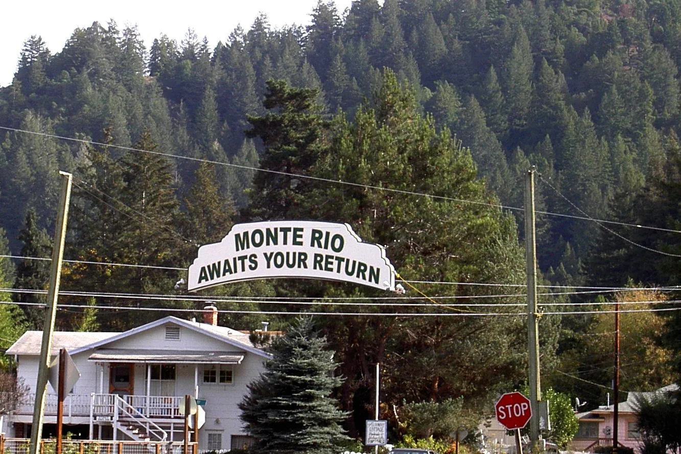 Arched Monte Rio Awaits Your Return sign over the road with redwood hills behind