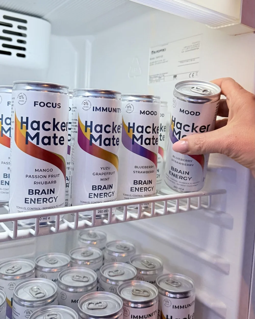 Nevera llena de latas de Hacker Mate de los tres sabores y una mano cogiendo una lata de Mood