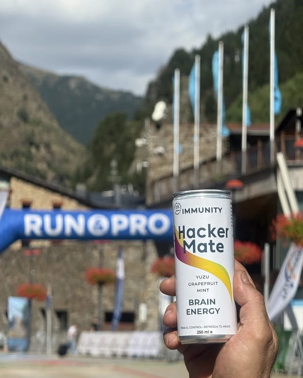 Mano sosteniendo una lata de Hacker Mate Immunity frente a la meta de una carrera en Andorra