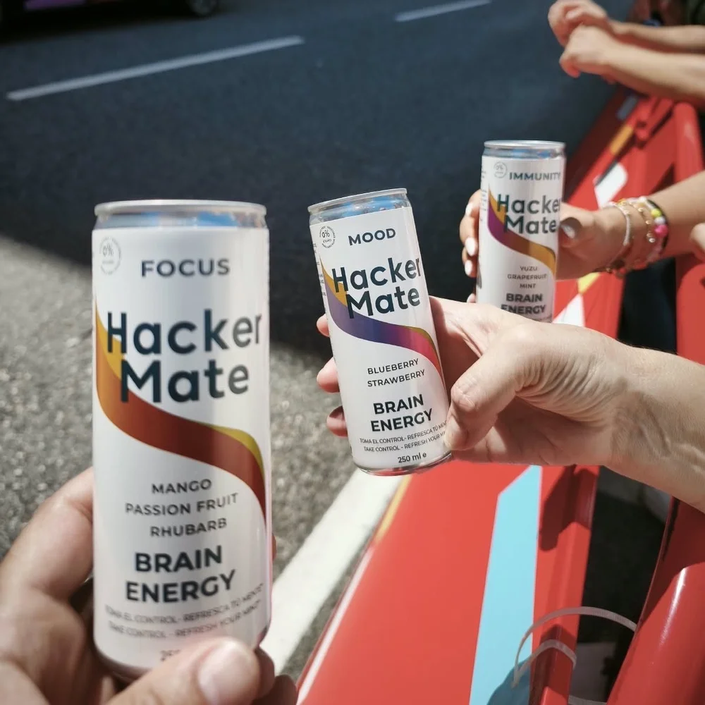Tres manos sostienen latas de Hacker Mate Focus, Mood e Immunity junto a una carretera durante una carrera