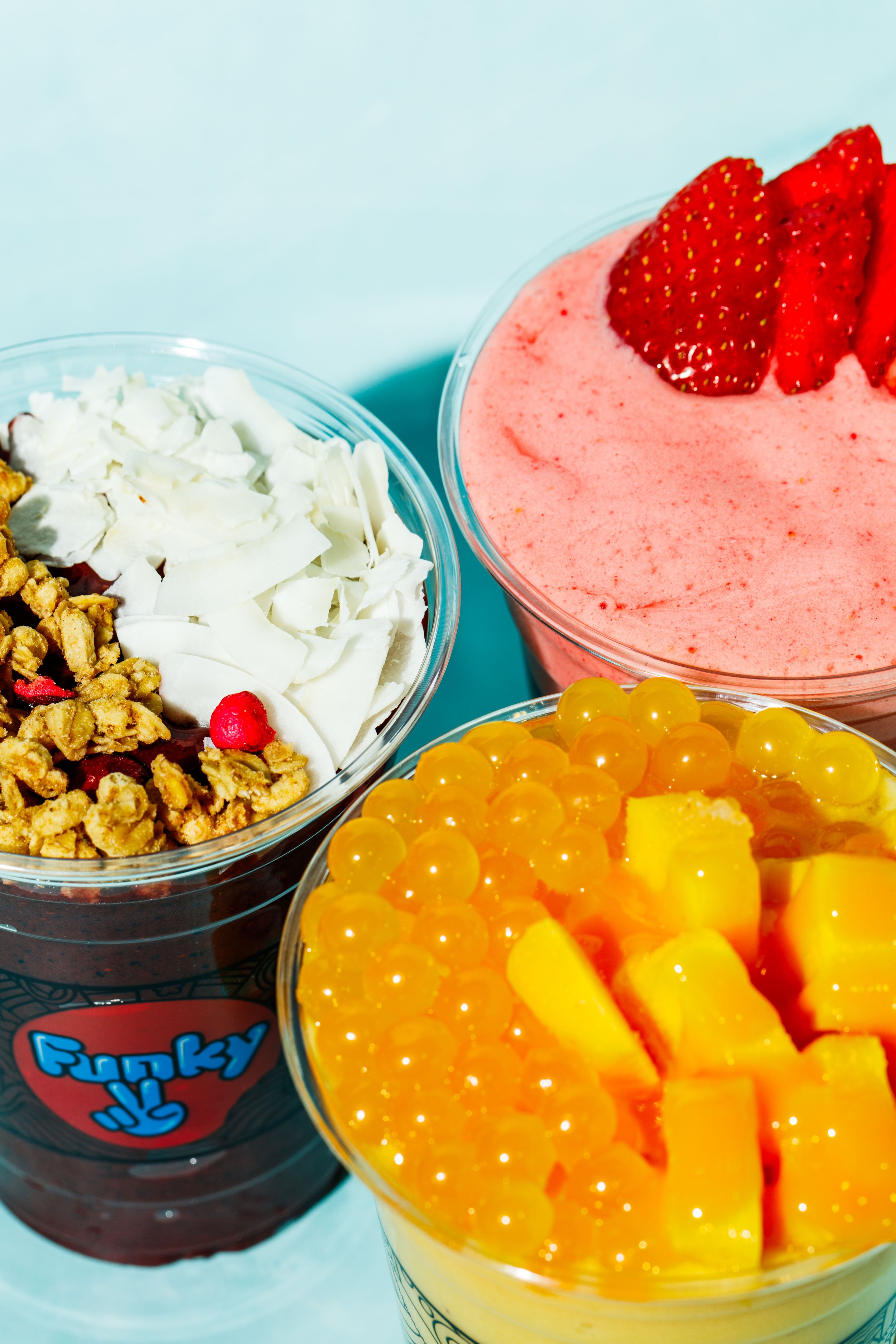 Tre kopper frossen yoghurt for Funky Frozen: açaí med granola og kokosflak, jordbær med ferske jordbær og mango med sprøe perler, på lysblå bakgrunn.