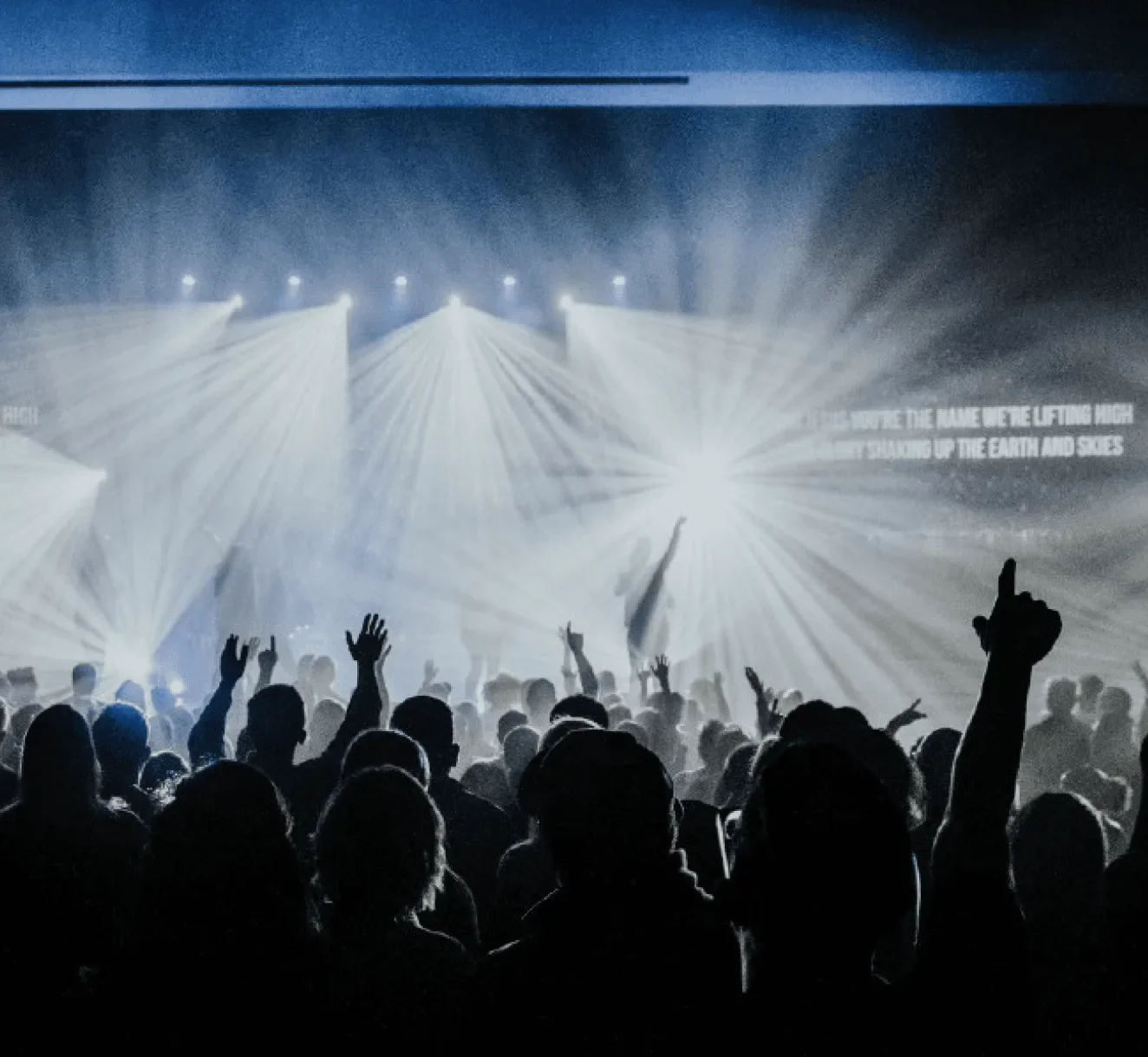 A crowd with hands raised in a large worship service under bright stage lights