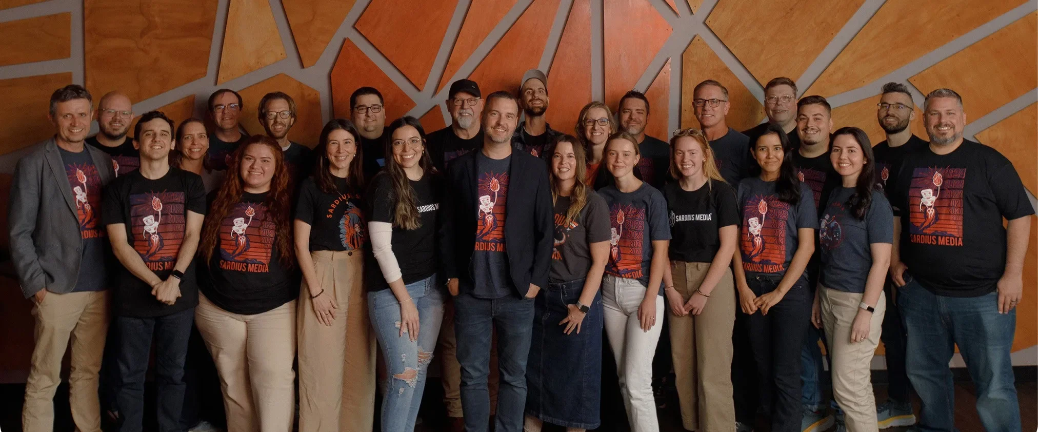 The Sardius Media team posed together in matching t-shirts in front of a warm, geometric wall