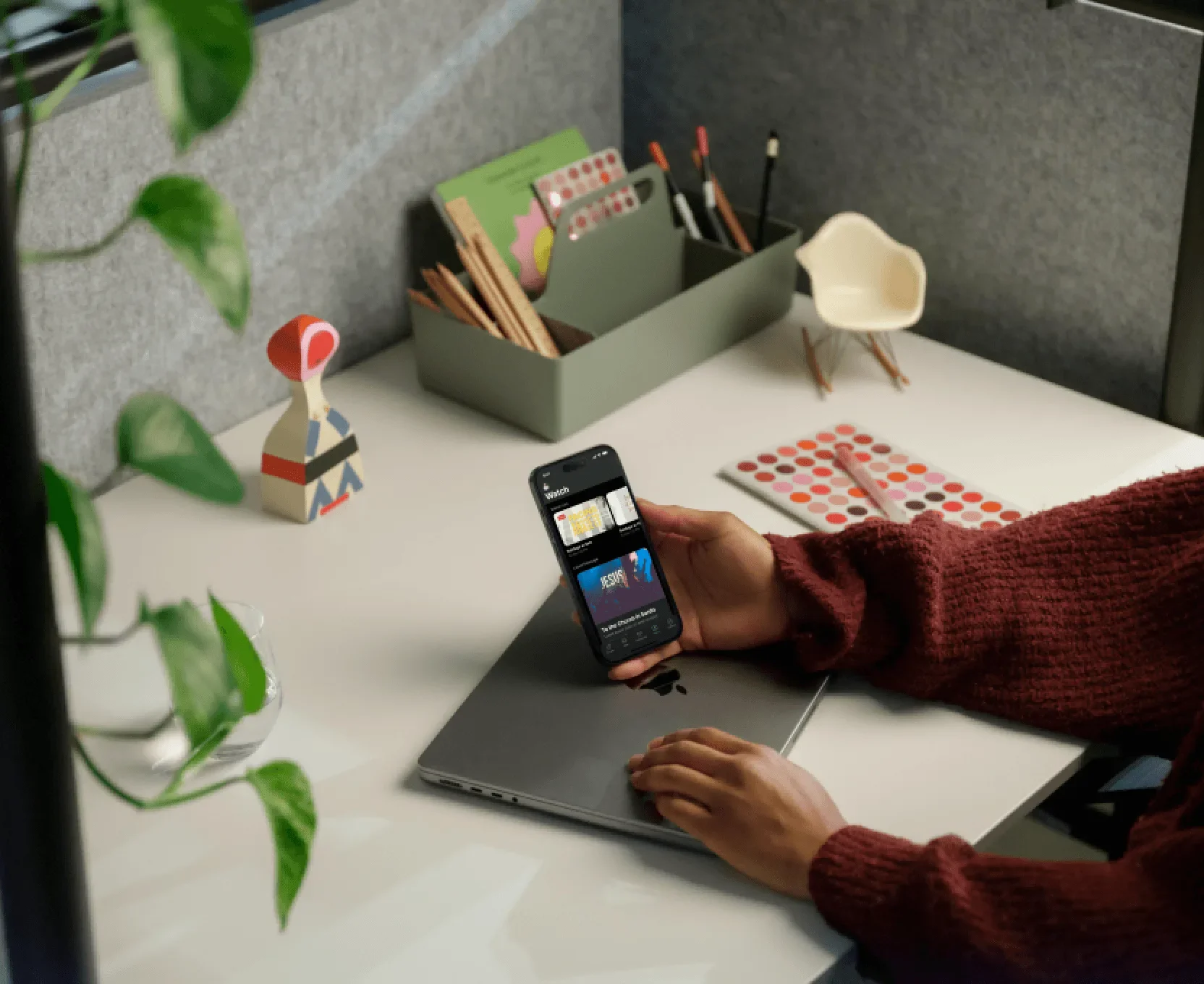 A hand holding a phone running a church app over a laptop on a tidy desk with plants