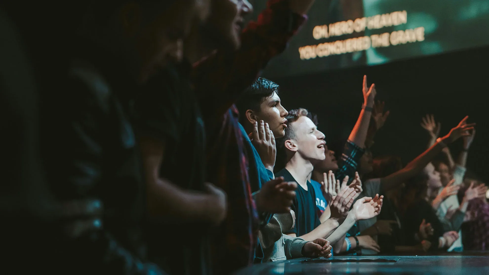 Young adults worshiping with hands raised in a dim, crowded room