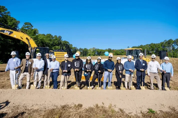 A church team in hard hats with shovels at a groundbreaking, an excavator behind them