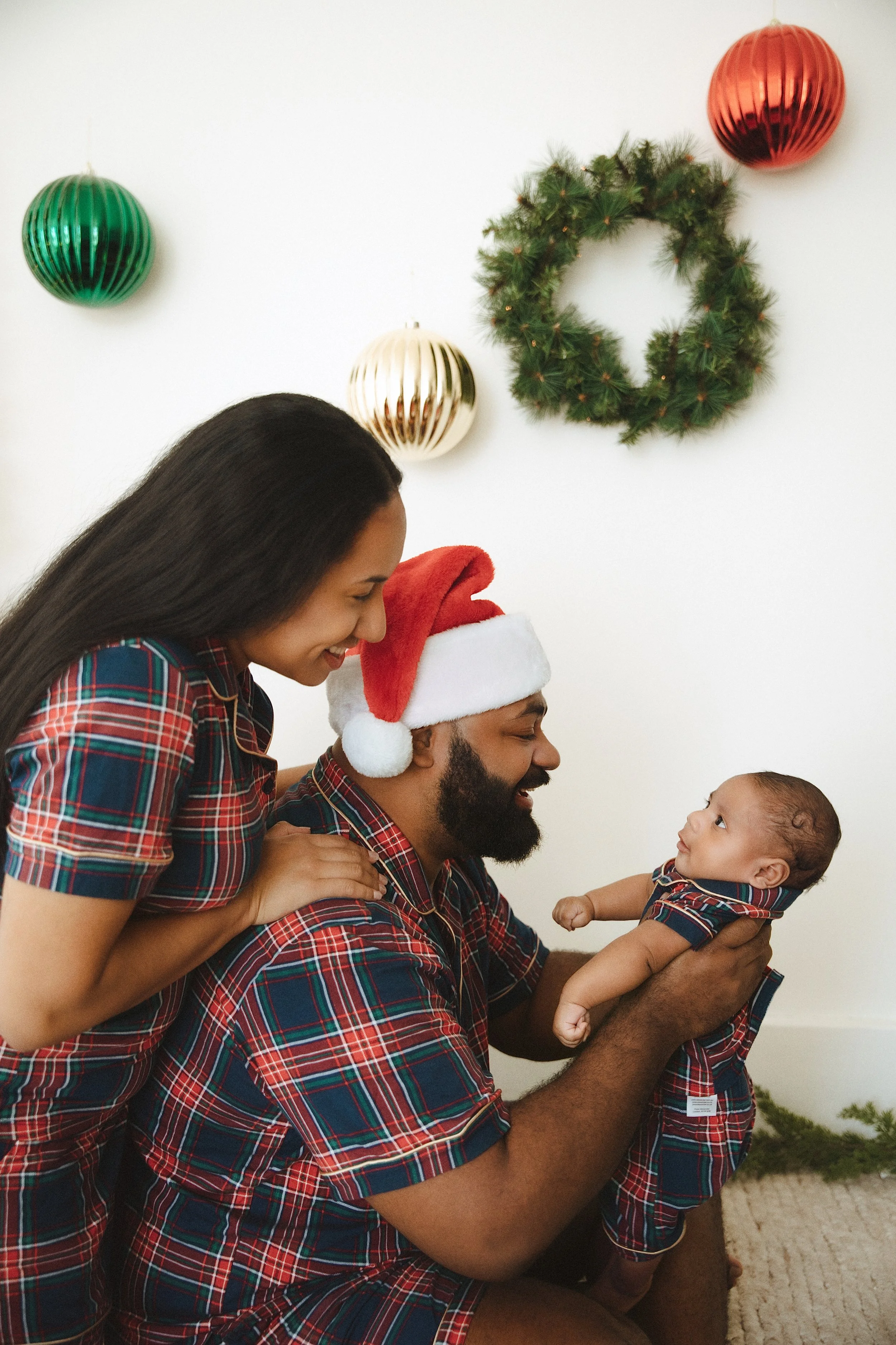 A mum and dad in matching plaid Christmas pyjamas smiling at their baby in a Foxie Studio Christmas session