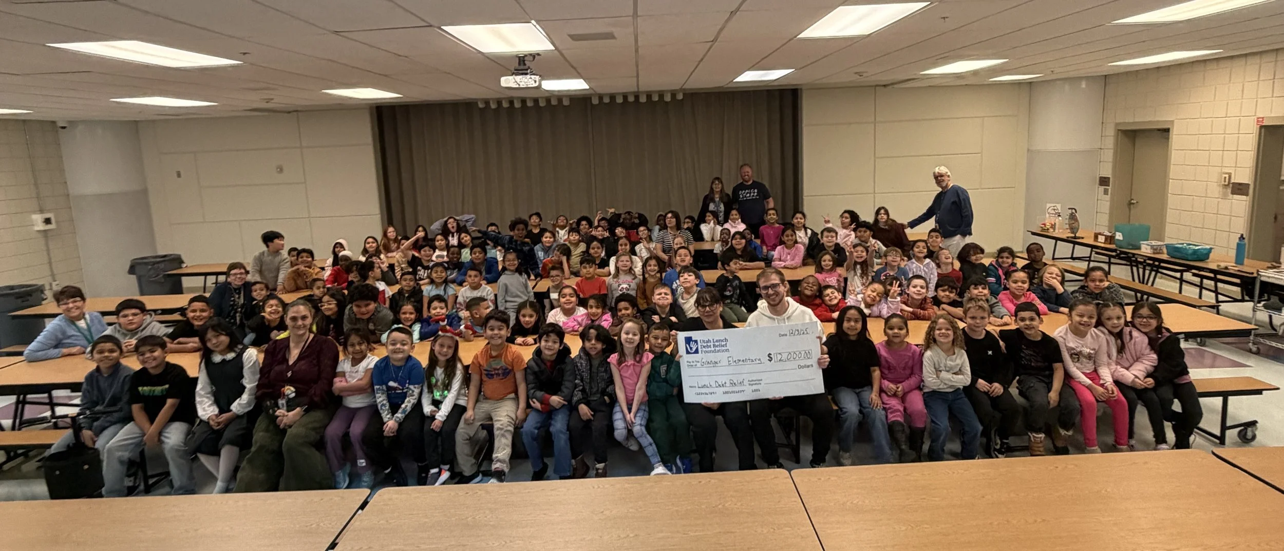 Founder in an elementary school cafeteria with a large group of students holding a Utah Lunch Debt Relief Foundation check.