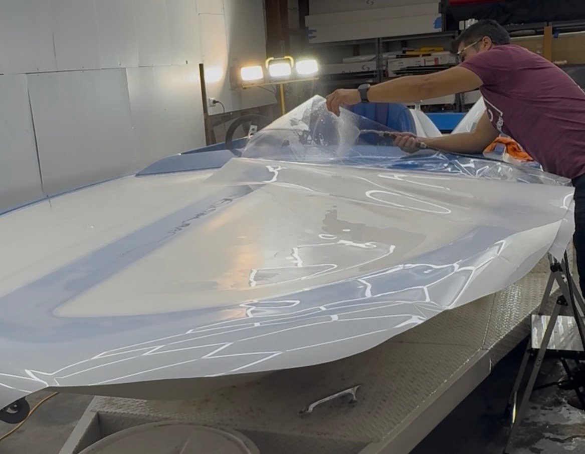 Installer positioning clear paint protection film across the blue-and-white boat’s top deck at RD studio.