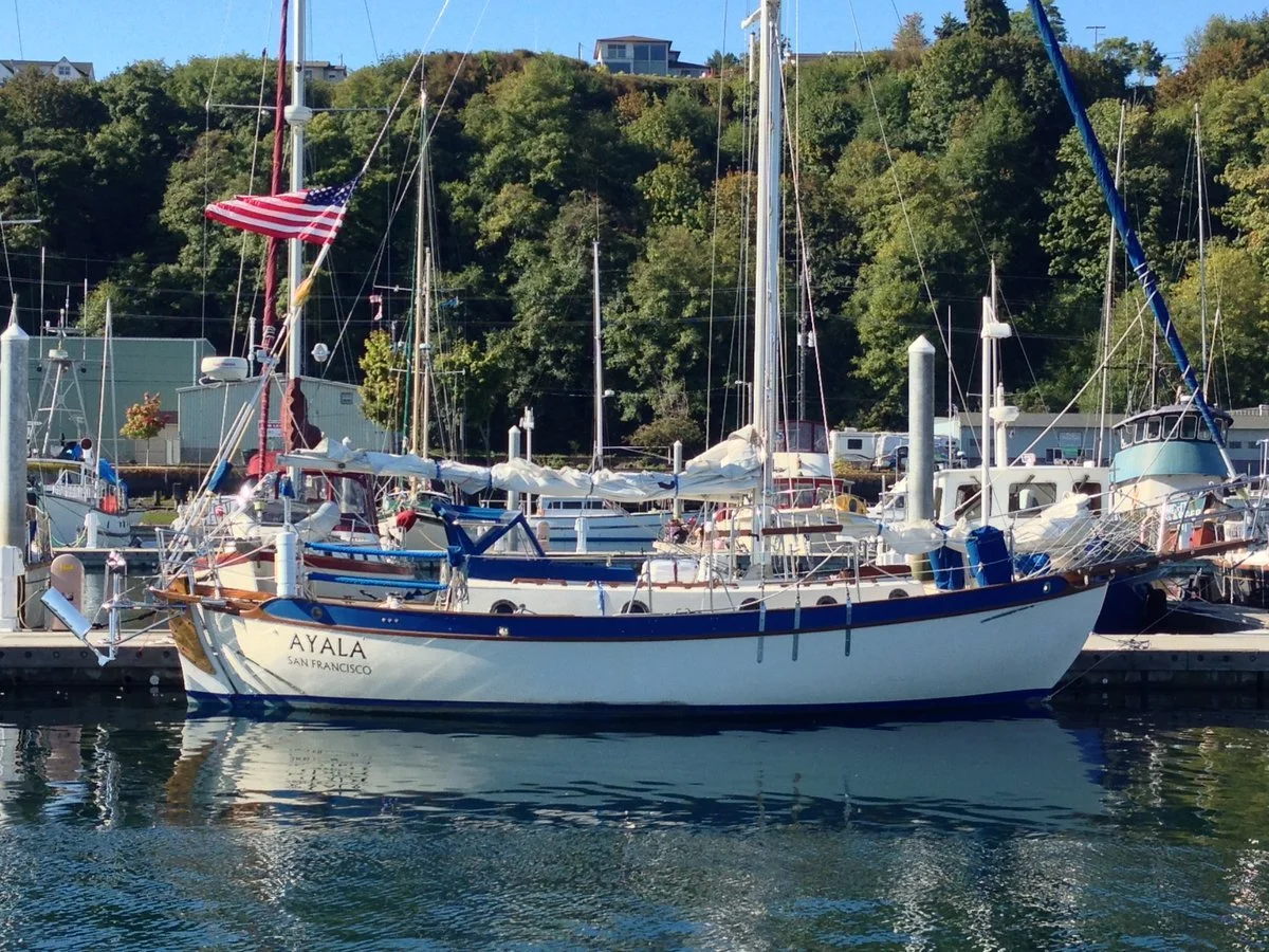 The sailboat Ayala tied up at a marina in Port Angeles, with a wooded hillside behind it.