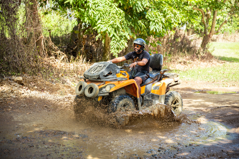A man rides an ATV through a muddy puddle splashing water up as he casually cruises through