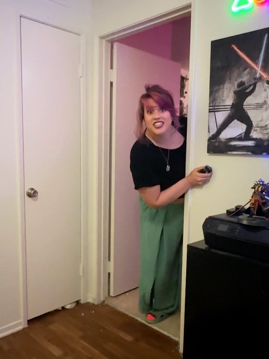 Woman peeking dramatically around a doorway with an excited expression in a gamer-themed room featuring Star Wars wall art and neon gaming lights.
