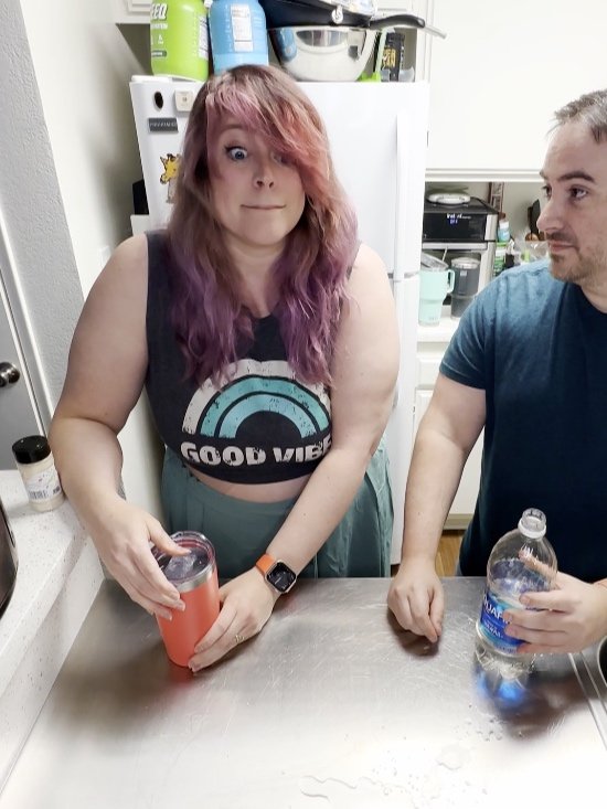 Woman with purple hair making a wide-eyed face while holding a pink drink next to man—funny couple moment.