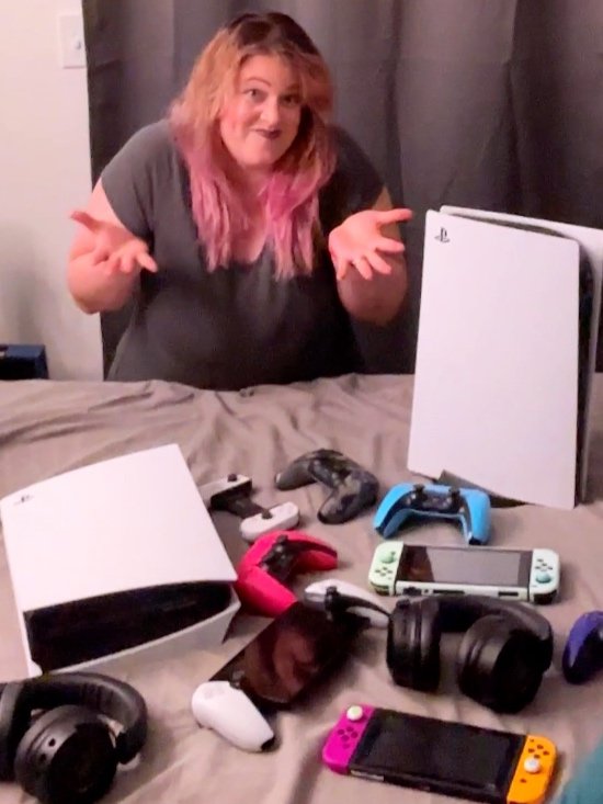 Woman shrugging dramatically behind a bed full of gaming gear including two PS5s, multiple controllers, headsets, and Nintendo Switch consoles—funny gamer overload moment.