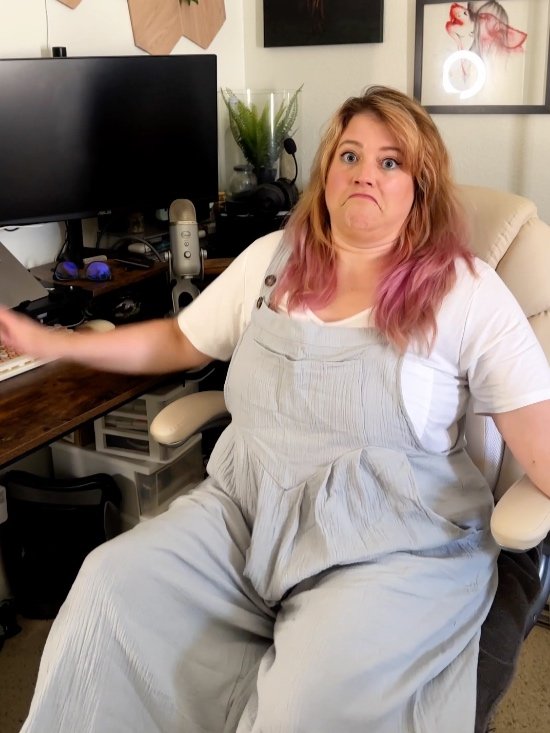 Woman with pink-tinted hair making dramatic face at home office desk—funny gamer reaction.