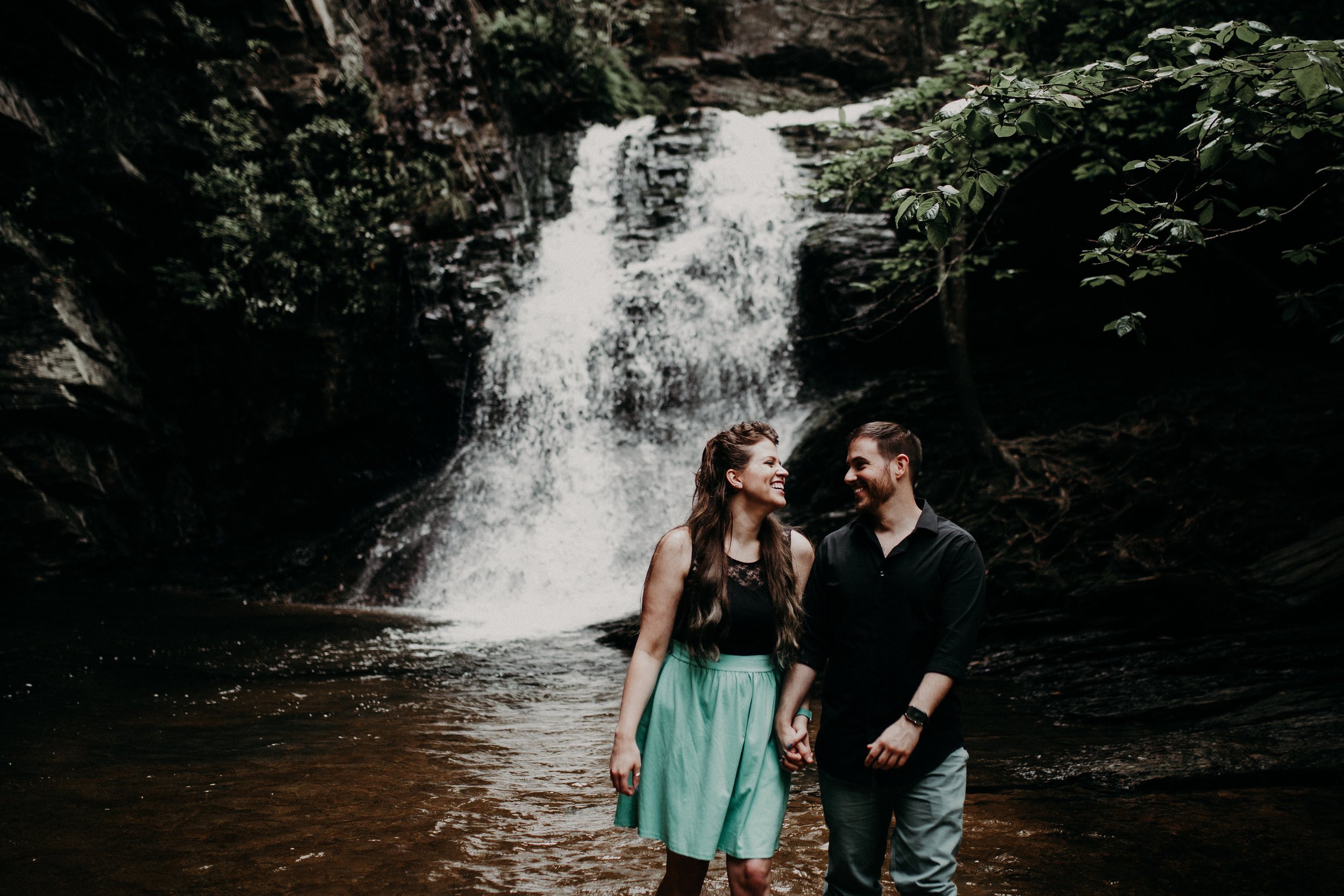 Robert and Michelle holding hands and smiling in front of a waterfall, playful outdoor couples photo for himVher lifestyle brand