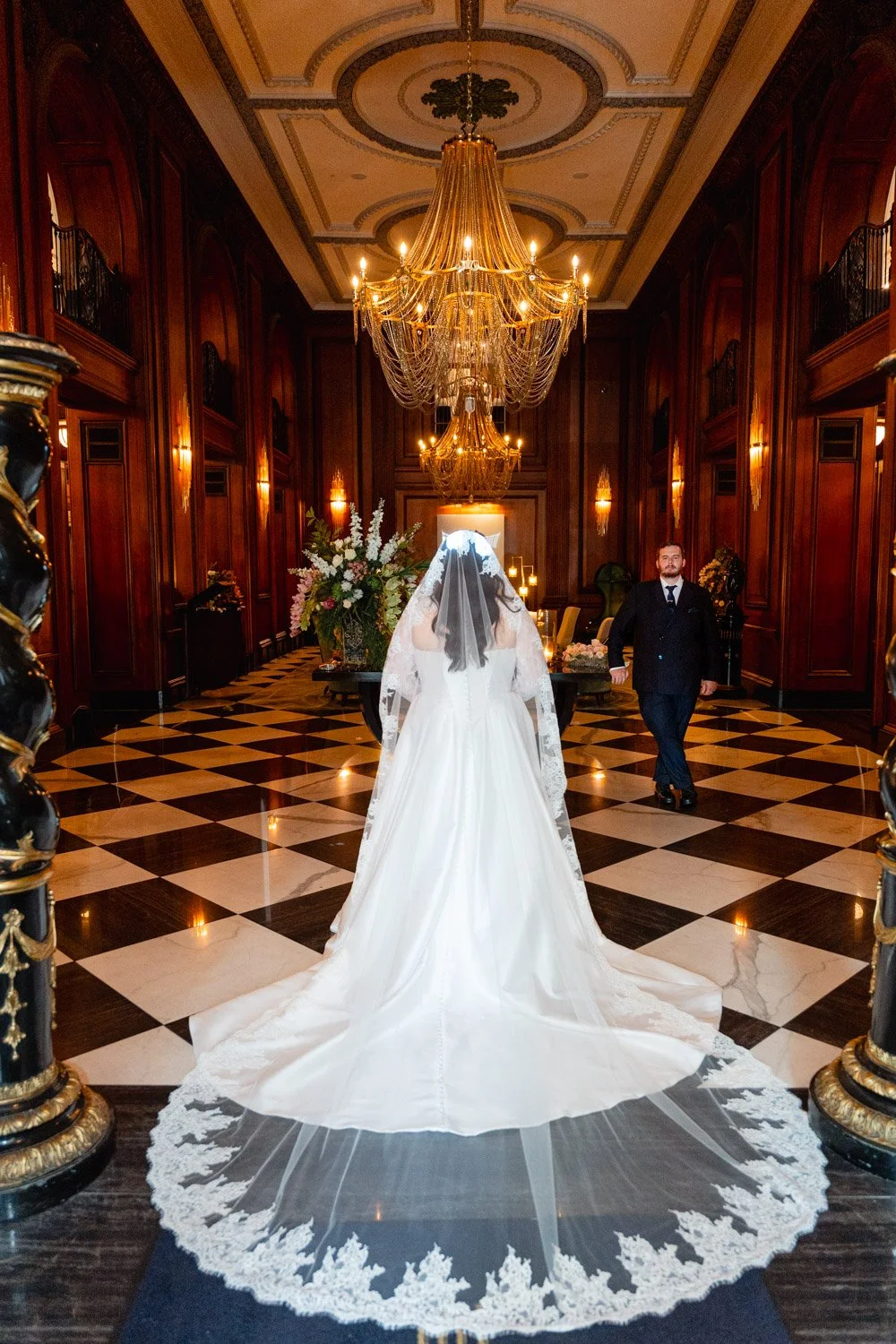 Bride with a long lace veil and groom beneath chandeliers