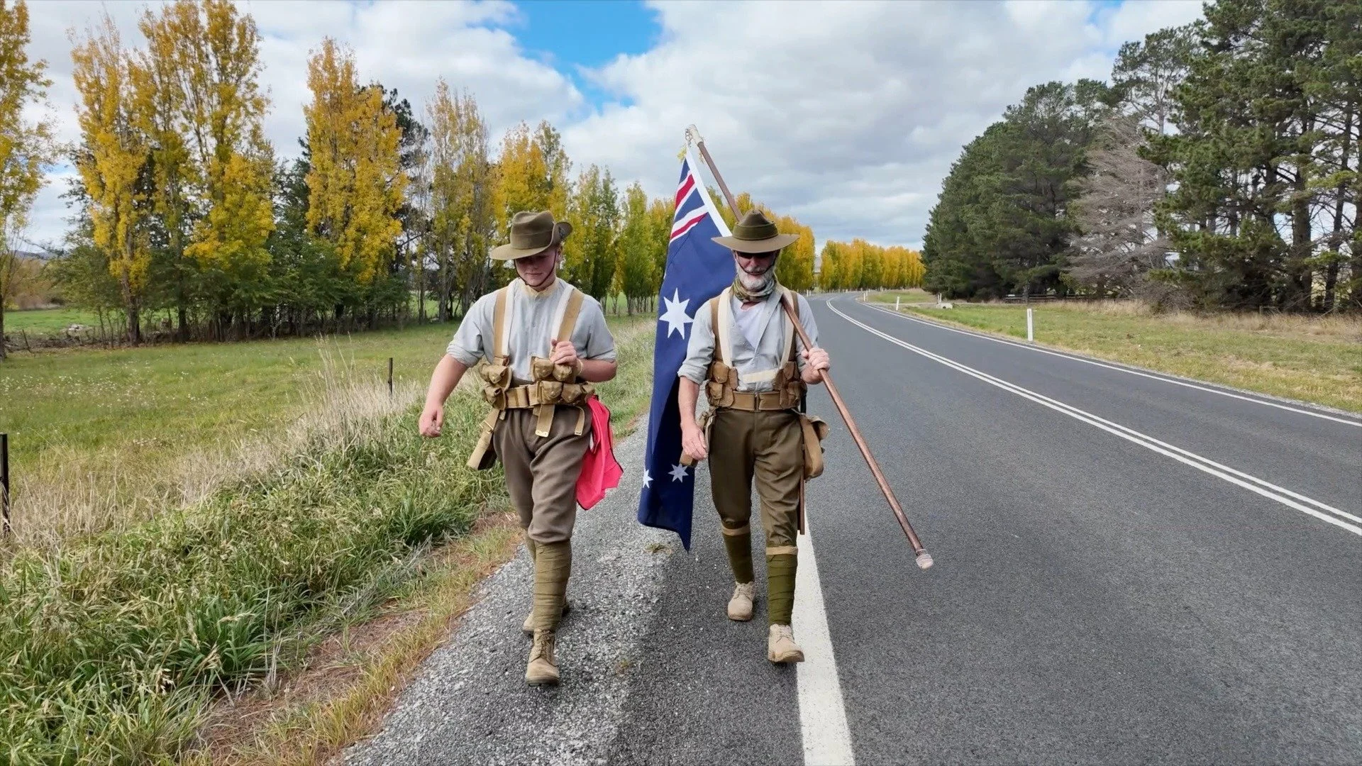 Two marchers walk the highway flanked by golden autumn trees during the Unknown Veteran March 2026
