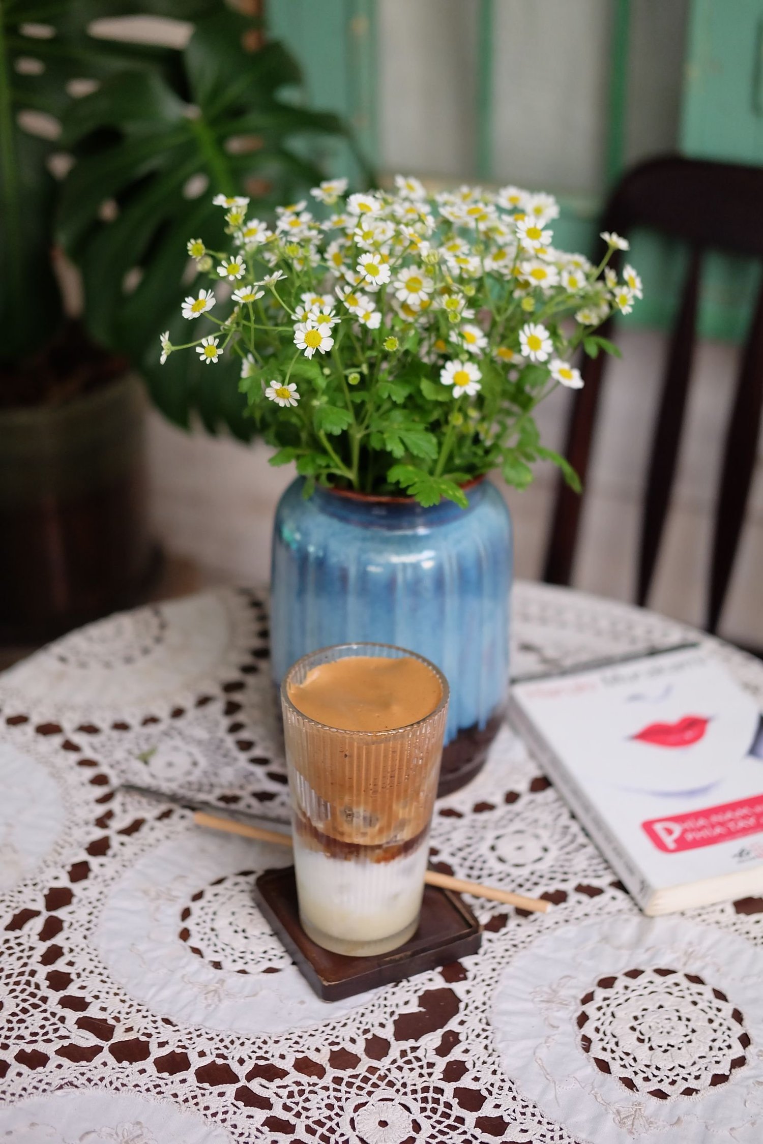 A glass of coffee beside a vase of daisies on a lace-covered table