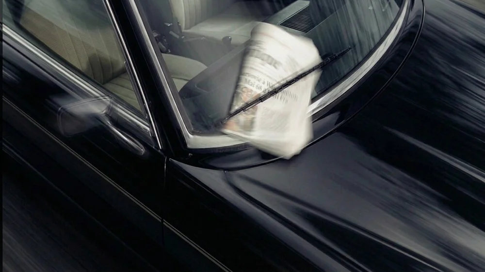 A folded newspaper caught under the windscreen wiper of a black classic car in motion, for BCLR's luxury print design service